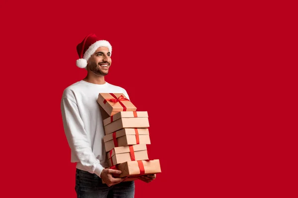 一个英俊的阿拉伯男子 戴着一堆堆礼物 手拿礼盒 孤零零地站在红色的背景下 快乐的中东青年男子背着圣诞礼物 遥望着旁边的复制空间 — 图库照片