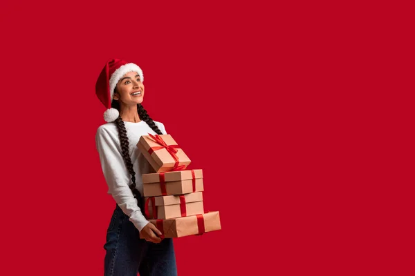 美丽的阿拉伯女人在圣诞老人的帽子里 手里拿着一堆堆礼物盒 孤零零地站在红色的背景下 快乐的年轻的中东女人背着圣诞礼物 遥望着复制的空间 — 图库照片