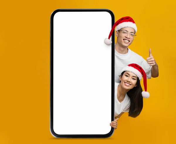 Happy chinese young woman and guy in Santa Claus hats show thumb up with big smartphone with blank screen isolated on yellow background. Offer, ad and app, advice for holiday, New Year and Christmas