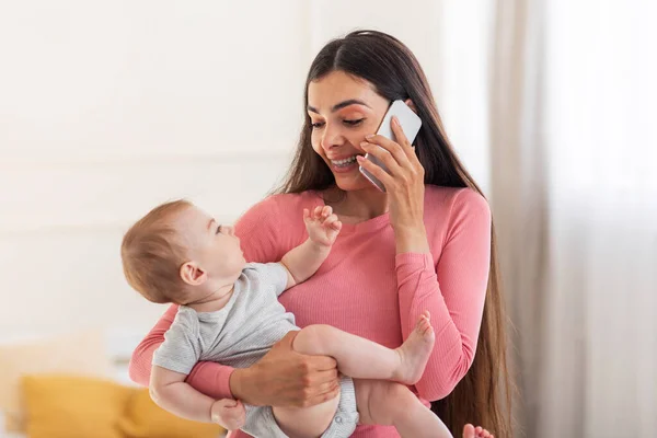 Young mothers lifestyle concept. Happy mom talking on cellphone, holding baby in arms and having phone conversation, sitting in bedroom interior at home