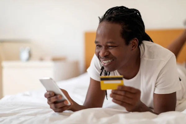 モバイル決済 スマートフォンやクレジットカードを手に黒の男を笑顔自宅でベッドでリラックス オンラインバンキングを使用して幸せな若いアフリカ系アメリカ人の男 電子商取引を楽しんで スペースをコピー — ストック写真