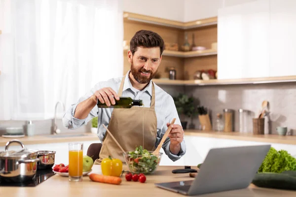 一个快乐的欧洲大胡子男人穿着围裙 把油倒在现代厨房的沙拉里 里面有有机蔬菜和笔记本电脑 在家里做健康午餐的新菜谱 在家里做饮食和食物博客 — 图库照片