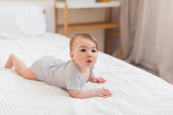 育児の概念 自宅の白いベッドシーツの上に横たわるかわいい赤ちゃんの身に着けている体のスーツの肖像画 フリースペース 快適な幼児はベッドルームでベッドの上でクロール — ストック写真