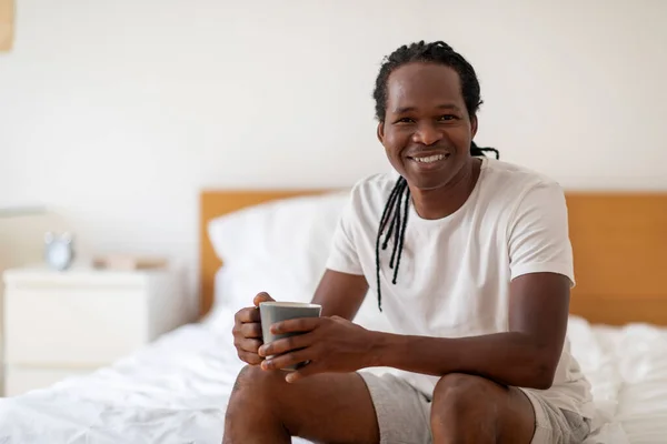 自宅で居心地の良いベッドルームでリラックスしながら 手でコーヒーを飲みながら ハンサムな若い黒人男性 千年紀のアフリカ系アメリカ人の男は カメラで熱い飲み物と笑顔を楽しんで スペースをコピー — ストック写真