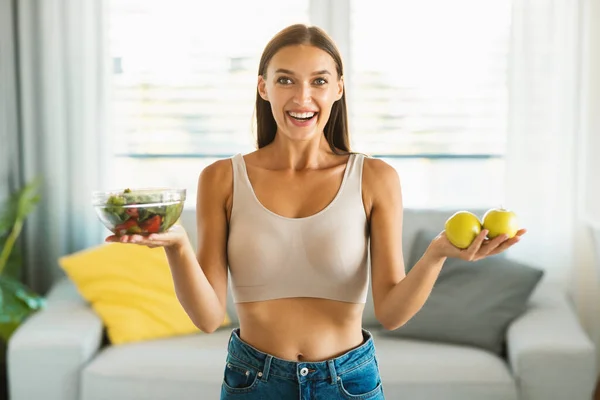 Healthy diet concept. Happy fit lady showing salad in bowl and apples, recommending weight loss nutrition, standing at home. Slimming concept