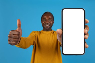 Neşeli orta yaşlı Afrikalı Amerikalı adam beyaz ekran ve başparmağıyla yepyeni bir akıllı telefon gösteriyor, en yeni mobil uygulamayı tavsiye ediyor, mavi stüdyo geçmişi, modelleme, kapatma