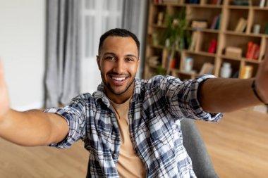 Heyecanlı Arap adam selfie çekiyor ve cep telefonu kamerasına gülümsüyor, evdeki koltukta oturuyor, boş yer var. Pozitif Orta Doğulu bir adamın portresi. Kapalı alanda kendi fotoğrafını çekiyor.