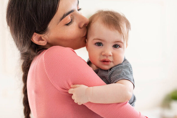 Caring young mother holding and kissing sweet little baby girl, mom and adorable kid bonding at home, copy space. Motherhood, moms love concept