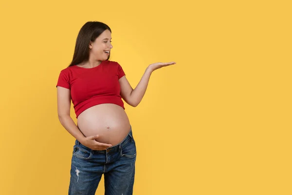 黄色の背景の上に立っている間 彼女のヤシの上に見えないオブジェクトを脇に見て若い妊婦の笑顔 デザインのためのコピースペースを示す陽気な期待の女性 — ストック写真
