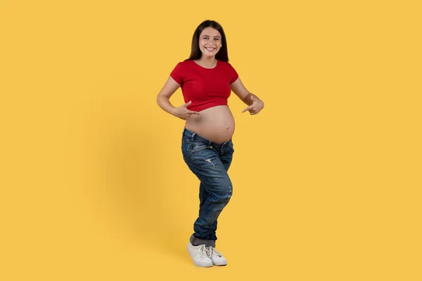 祝你怀孕愉快快乐的孕妇用两只手指著她的大美女 快乐的微笑着期待着的女士站在黄色的背景下展示她的腹部 复制空间 — 图库照片