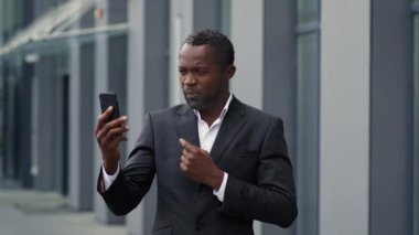 Çevrimiçi iş görüşmeleri. Profesyonel orta yaşlı siyahi iş adamı, ortaklarıyla akıllı telefon aracılığıyla konuşuyor, proje detaylarını açık havada tartışıyor, yavaş çekim, serbest alan