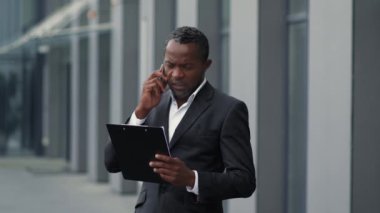Orta yaşlı Afro-Amerikan erkek menajer ortaklarla telefonda konuşuyor, iş planlarını tartışıyor, belgelerle pano tutuyor, ofis binasının yanında duruyor, yavaş çekim, boş alan
