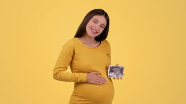 若いです幸せな妊娠中の女性保持Sonography写真の彼女の赤ちゃんと笑顔 愛撫とTickling彼女の大きな腹 黄色のスタジオの背景 遅い動き — ストック動画