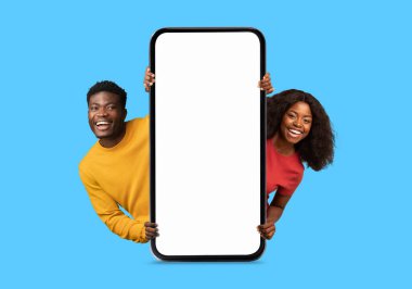 Gülümseyen Afro-Amerikan milenyum çifti boş ekranlı büyük bir akıllı telefondan dışarı bakıyor, blog tavsiye ediyor, mavi arka planda, stüdyoda izole edilmiş. Online teklif, reklam ve uygulama, satış ve yüz ifadesi