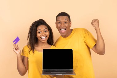 Heyecanlı siyah çift boş dizüstü bilgisayar ekranı gösteriyor, kadın kredi kartı tutuyor ve birbirine yumruk sallıyor, tasarım için maket yapıyorlar. Genç adam ve kadın online alışveriş yapıyor.