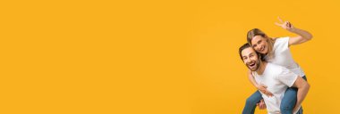 Pozitif genç çift kamerada barışı sağlarken ve eğlenirken uzun süre mutlu sevgilisini sırtında taşıyan neşeli adamın sarı arka planda, geniş yatay pankartta poz verirken görüntüsü...