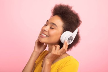 Neşeli, rahat, bin yıllık Afro-Amerikan kıvırcık saçlı sıradan, kablosuz kulaklıklı bayan pembe arka planda, stüdyoda izole edilmiş gözlerle müziğin tadını çıkarıyor. İnsani duygular, boş zamanlarında dinlenmek için ses uygulaması