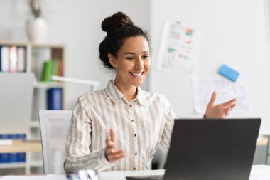 Heyecanlı kadın girişimci videosu dizüstü bilgisayarla arıyor, web kamerasıyla konuşuyor, gülümsüyor, internette iletişim kuruyor, iş yerinde oturuyor.