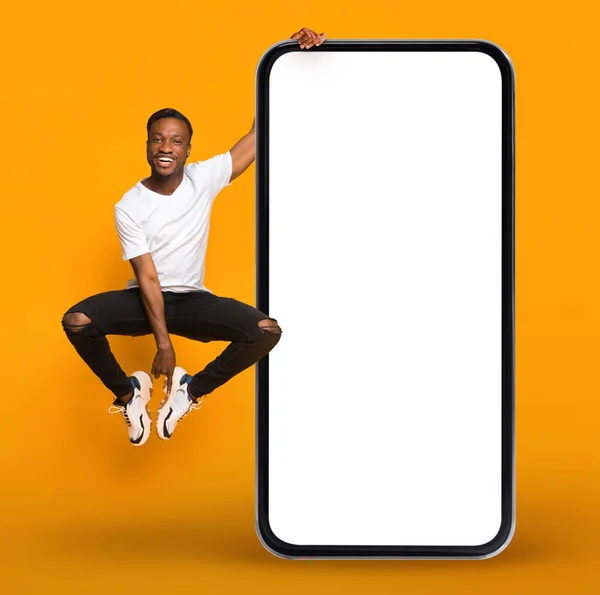 快乐的年轻黑人在巨大的空白手机旁边跳着 屏幕是白色的 兴奋的非洲裔美国人背景是橙色的 展示了手机广告的复制空间 — 图库照片