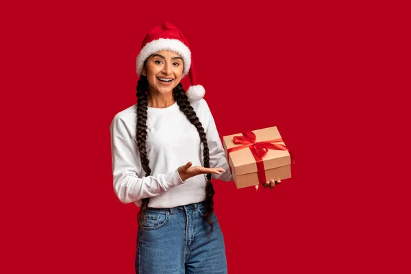 假日奖金 漂亮的阿拉伯女人在圣诞帽的包装礼品盒里 快乐的年轻中东女性一边手牵着手 一边翻阅红色的工作室背景 复制空间 — 图库照片