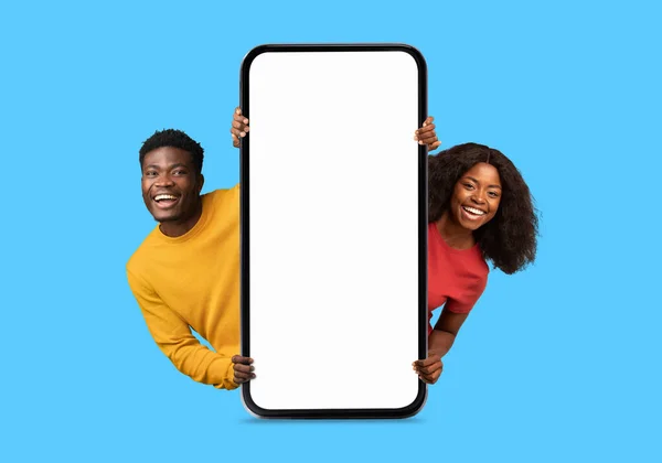 笑着的非洲裔美国千禧年夫妇从巨大的智能手机上用空屏幕偷窥 推荐博客 隔离在蓝色背景下 工作室 在线提供 广告和应用 销售和面部表情 — 图库照片