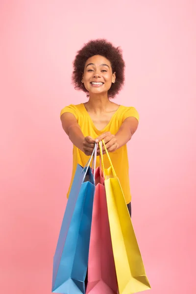 黄色のTシャツの陽気な千年紀のアフリカ系アメリカ人の巻き女性の学生は ピンクの背景 スタジオ 垂直上に隔離された購入と多くのバッグを示しています オンラインショッピング 巨大な販売 広告やオファー — ストック写真
