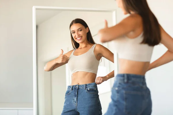 幸せな若い女性がジーンズに身を包み 新しいサイズを承認し 親指をジェスチャーし 笑顔で 鏡の近くに立って 自由空間 私は体が好きだ — ストック写真