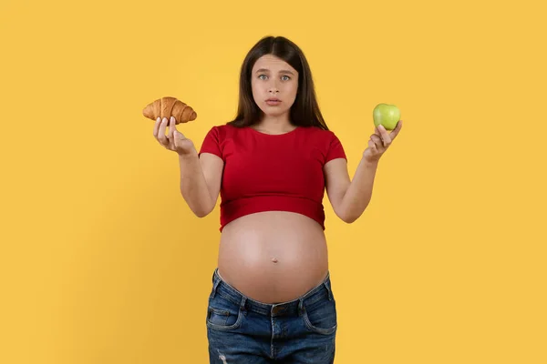 妊娠中の女性が黄色の背景の上に立っている間に健康的で健康的でない食べ物の間で選択している疑いのある若い期待の女性を手に入れ クロワッサンと緑のアップルを保持混乱 スペースをコピー — ストック写真