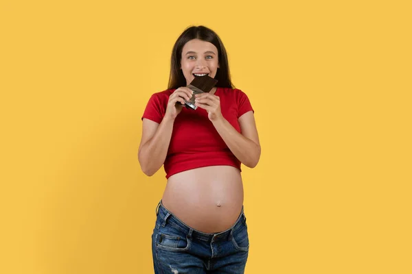 怀孕的渴望 快乐的年轻怀孕妇女 边吃巧克力条边站在黄色的背景上看着相机 愉快地期待着女性吃糖果当点心 复制空间 — 图库照片