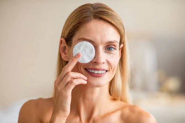 Facial skincare. Beautiful middle aged lady covering eye with cotton pad, posing and smiling to camera. Beauty and skin care cosmetics concept