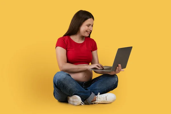 オンラインショッピング 黄色のスタジオの背景の上に床に座っている間 ラップトップコンピュータを使用して幸せな若い妊娠中の女性 インターネットで購入する期待の女性を笑顔 フリースペース — ストック写真