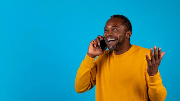 刺激的なニュースだ 感情的にハンサムな成熟した黒人男性で黄色の携帯電話の会話 ジェスチャー 広告のためのコピースペースを見て 青いスタジオの背景に隔離された パノラマ — ストック写真
