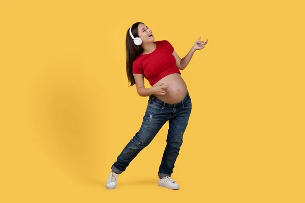 ワイヤレスヘッドフォンで音楽を聞き エアギターを演奏する正の若い妊婦 スタジオで黄色の背景の上に立って楽しみを持っている陽気な無料の期待の女性 スペースをコピー — ストック写真