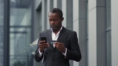Modern mali işlemler. Başarılı orta yaşlı Afro-Amerikan iş adamı kredi kartı ve akıllı telefonuyla online işlem yapıyor, ofis binasının yanında duruyor, yavaş çekim, serbest alan