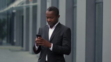 Modern iletişim. Ciddi bir orta yaşlı Afro-Amerikan iş adamının açık hava portresi. Akıllı telefondan mesajlaşıyor, ortaklarla sohbet ediyor, ofis binasının yanında duruyor, yavaş çekim, serbest alan.