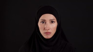 İslami kadınları kurtarın. Geleneksel başörtüsü takan üzgün Müslüman kadının yakın plan portresi. Kameraya ciddi ciddi bakıyor, siyah stüdyo arka planında poz veriyor, ağır çekim.