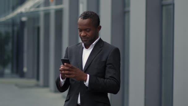 現代のコミュニケーション 深刻な中年アフリカ系アメリカ人ビジネスマンの屋外の肖像画スマートフォンでテキストメッセージ パートナーとチャット オフィスビルの近くに立って スローモーション フリースペース — ストック動画