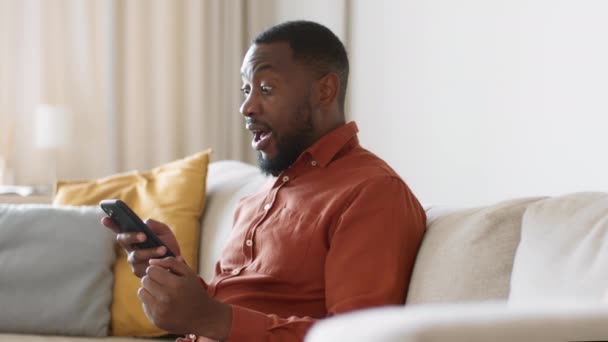 网上赢的概念 年轻的非洲裔美国人在智能手机上冲浪 获得了胜利的信息 庆祝了成功 在家里躺在沙发上 跟踪镜头 慢动作 自由自在 — 图库视频影像