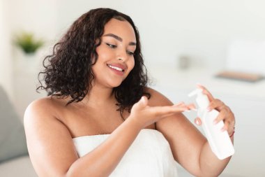 Mutlu Afrikalı Amerikalı tombul kadın elinde nemlendirici losyon sürüyor. Havluya sarılmış kadın yatak odasında oturuyor.