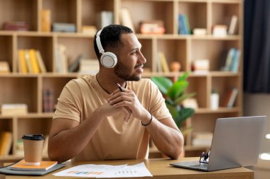 Evde çalışan, bilgisayar başında çalışan, kulaklık takan ve fotokopi odasına bakan düşünceli, düşünceli bir Arap.