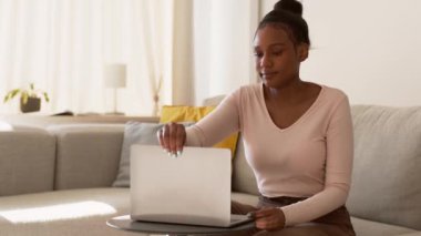 Konforlu serbest çalışma konsepti. Genç ve güzel Afrikalı Amerikalı kadın dizüstü bilgisayarı açıp yazmaya başlıyor, evden online olarak çalışıyor, çekim takip ediyor, yavaş çekim, boş alan