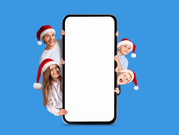 带着孩子戴着圣诞礼帽的欧洲裔年轻父母笑着从大智能手机里探出头来 屏幕空空荡荡 与蓝色背景 工作室隔离 新年庆祝活动 假日优惠 广告和网络应用 — 图库照片