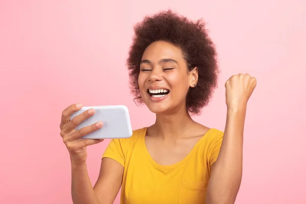 心满意足的年轻非洲裔美国卷曲女士在智能手机上读到信息 做出成功和胜利的手势 与粉色背景隔离 网上赢 大新闻 广告和报价 — 图库照片