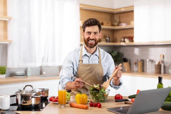在现代厨房用有机蔬菜和笔记本电脑准备沙拉的过程中 笑着的欧洲成年大胡子男性厨师在围裙 家里的食物博客 家里做健康晚餐的新菜谱和饮食 — 图库照片