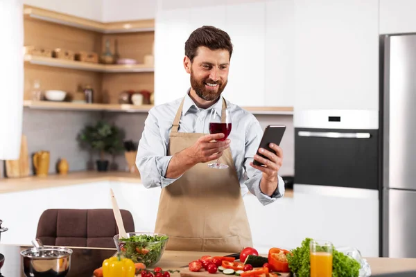 Happy adult european male in apron prepares dinner, holds glass of wine, looks at phone in kitchen interior with vegetables. Cook meal, food blog at home, holiday celebration remotely and video call