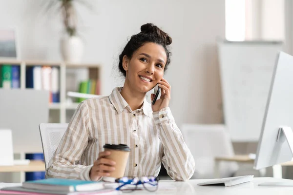 Happy female entrepreneur talking on cellphone and drinking coffee at workplace in office, smiling woman holding cup with takeaway drink and enjoying pleasant conversation