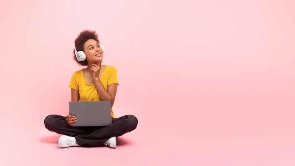 stock image Smiling pensive millennial african american curly female in wireless headphones, sit on floor with laptop, enjoy music, look at empty space, isolated on pink background, studio. Great ad and offer