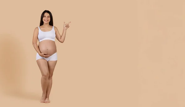 妊娠中の広告 コピースペースで脇を指差す下着姿の綺麗な妊婦さん スタジオでベージュの背景の上に立ちながらカメラを見ている幸せな若い期待の女性 パノラマ — ストック写真