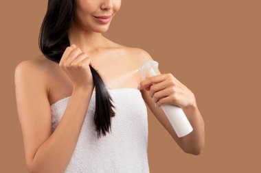 Tanımlanamayan, yarı çıplak, uzun saçlı, beyaz banyo havlusuna sarılı, duş üstüne saç spreyi kullanan, bej renkli stüdyo arka planında izole edilmiş, fotokopi çekilen esmer kadın. Saç şekillendirme konsepti