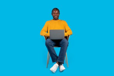 Sarı kazaklı mutlu Afrikalı orta yaşlı adam, mavi stüdyo arka planında modern laptop kullanıyor, klavyeye yazıp gülümsüyor, çalışıyor ya da oyun oynuyor, boşluğu kopyalıyor.